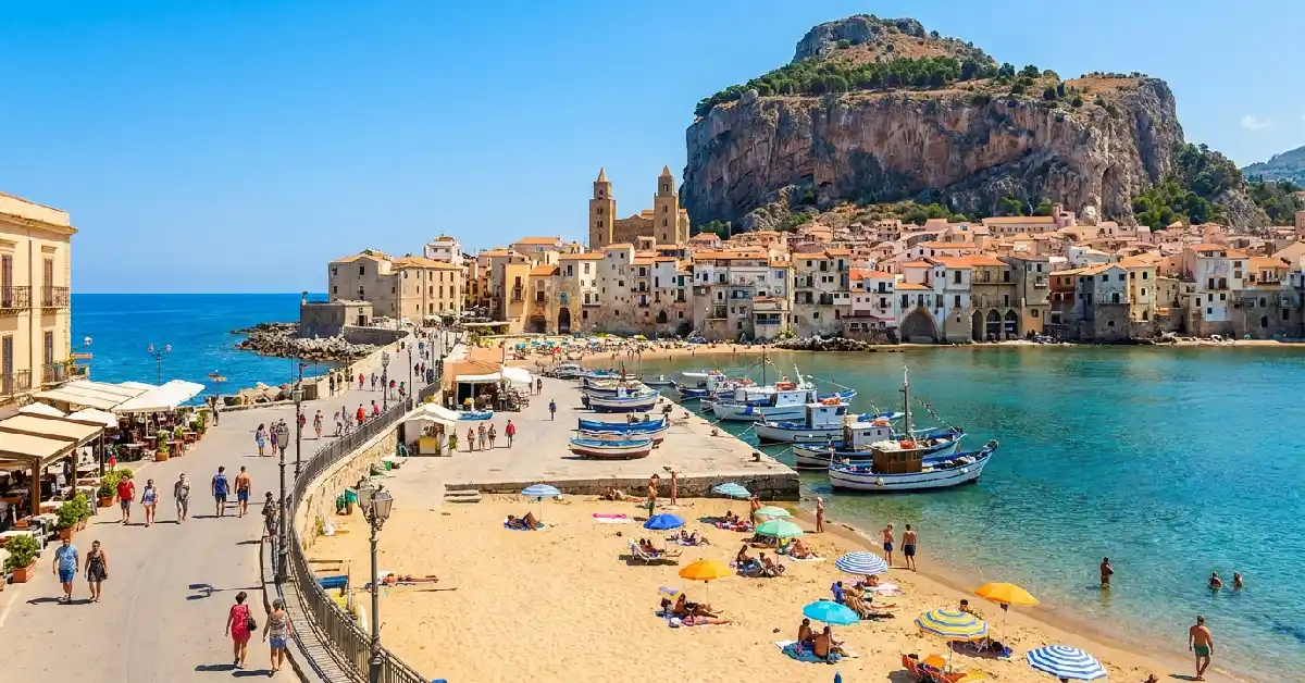 Cefalù golden beach and Norman cathedral along the Lungomare promenade in Sicily under bright sunlight.