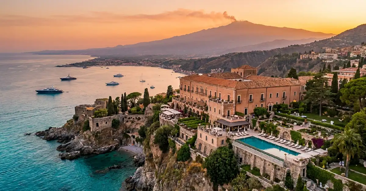 White Lotus Filming Locations Sicily Visit, Cinematic aerial view of San Domenico Palace on cliffs above the Ionian Sea in Taormina Sicily at sunset with Mount Etna in background.
